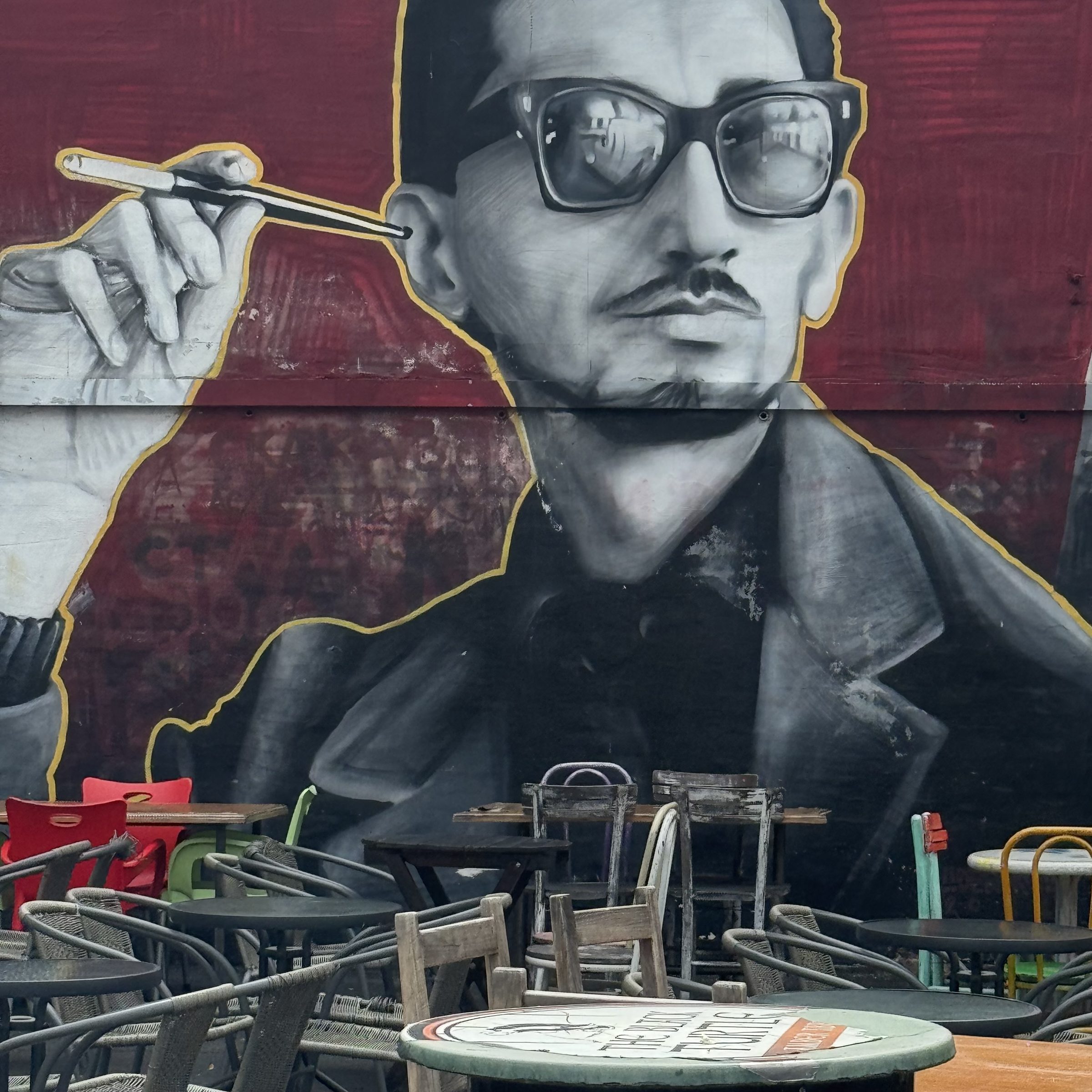 A large black-and-white street mural of a man in sunglasses holding a cigarette holder on a deep-red wall, above an outdoor café with stacked chairs in Belgrade.