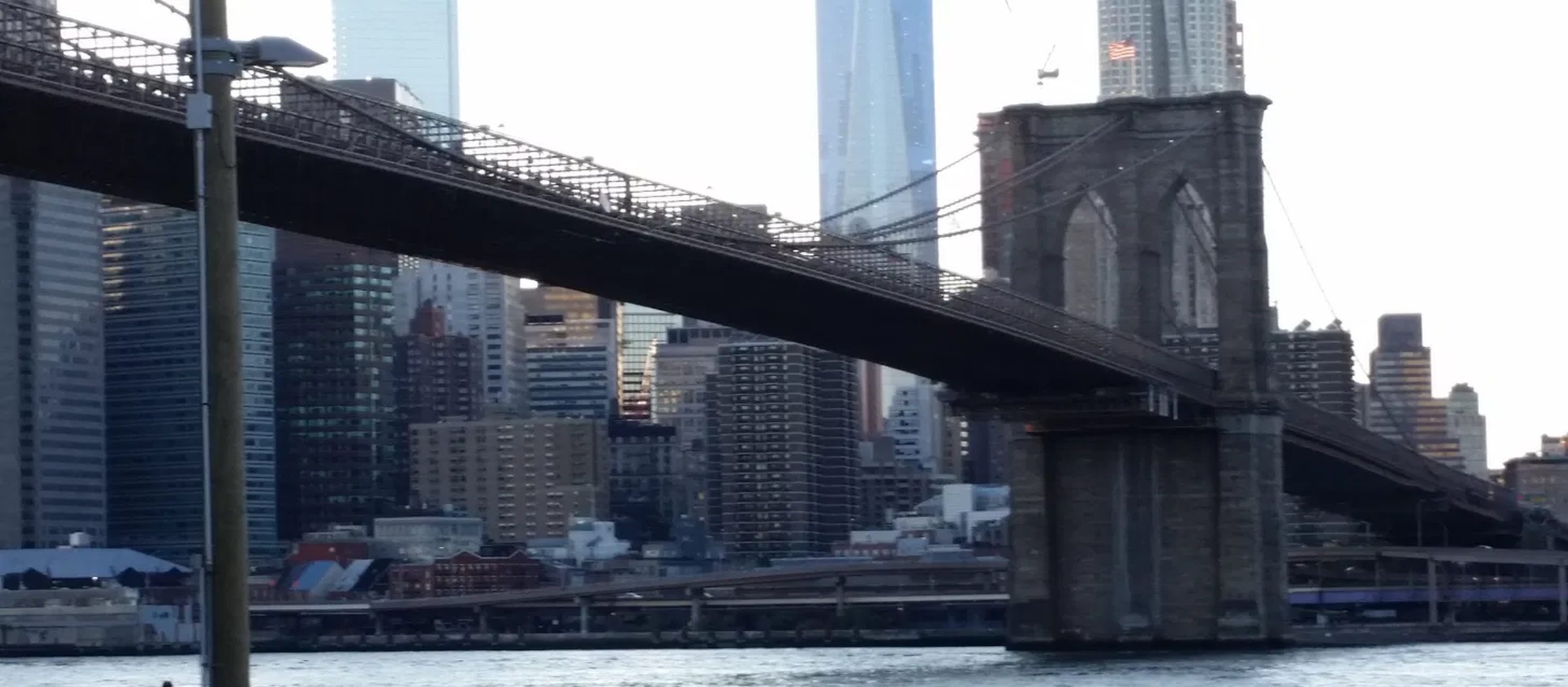 The Brooklyn Bridge spanning the East River with the Lower Manhattan skyline, including One World Trade Center, rising behind it.