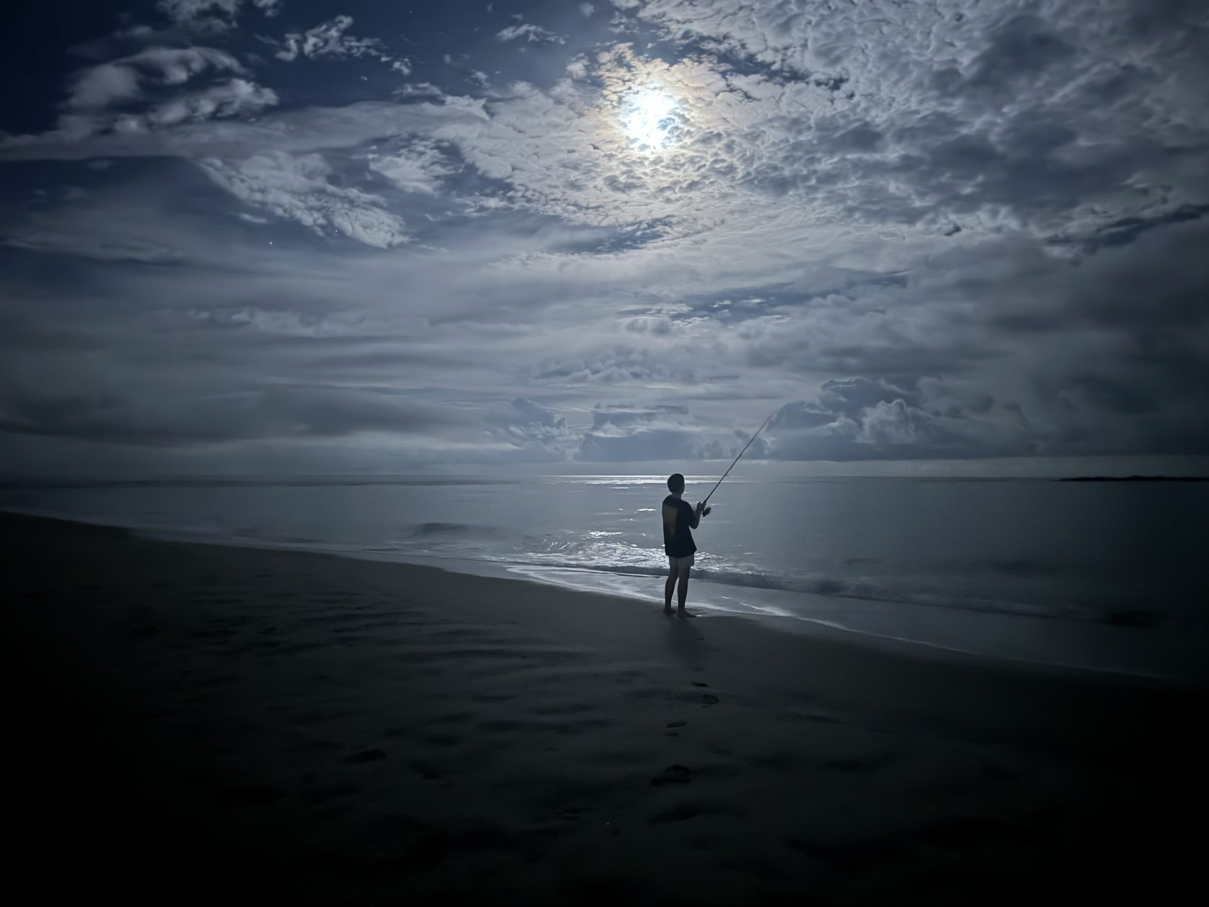 A lone angler casting a line into the surf on a moonlit beach at night, the moon breaking through clouds over the ocean.